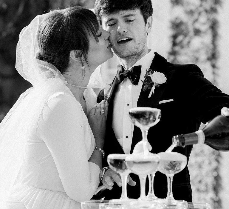 Bride kisses her groom on the cheek as he pours champagne into champagne tower on their wedding day