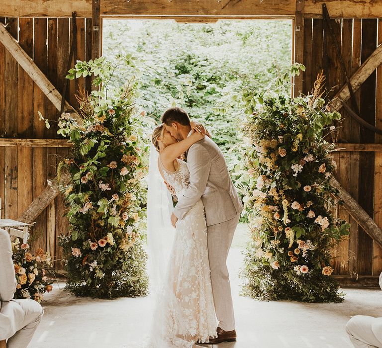 Bride and groom share their first kiss as a married couple in front of the flower column altar decorations at Nancarrow Farm