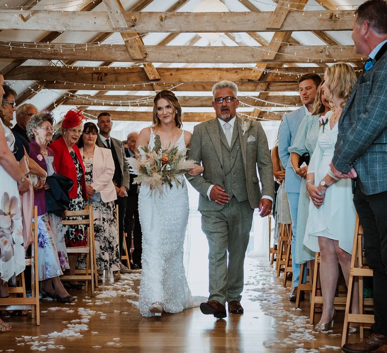 Bride in sleeveless Made With Love Bridal dress with white embellishments holding dried flower and burnt orange and toffee rose bouquet walking down the aisle with her dad in grey suit