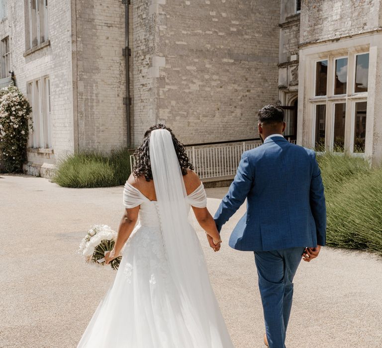 Bride wears off the shoulder wedding dress and cathedral veil whilst walking with her groom in blue suit 