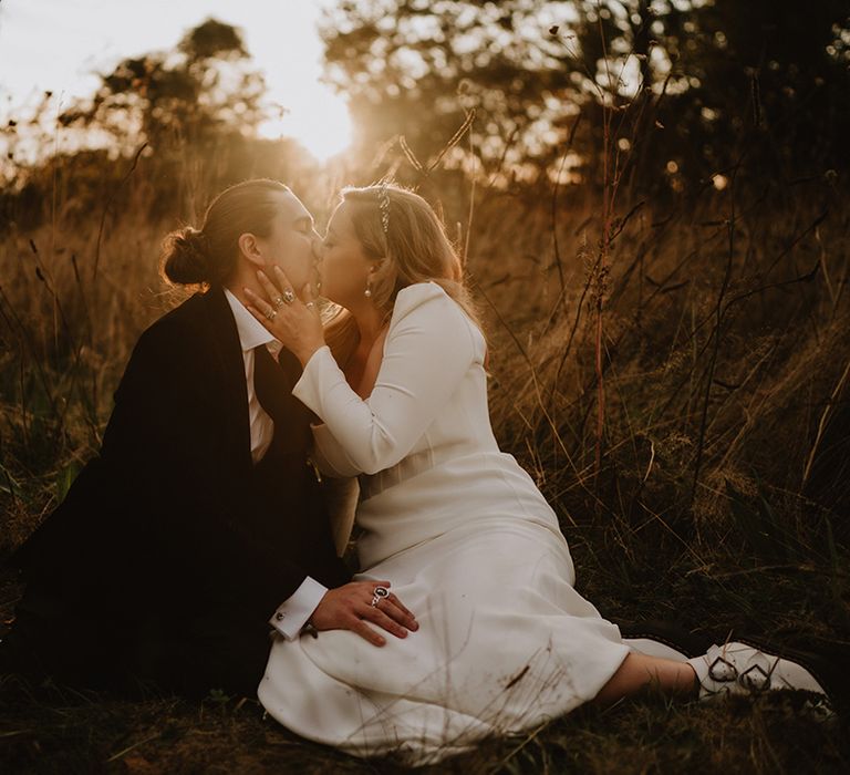 Bride in square neck long sleeve satin wedding dress with two tier veil sitting with and kissing groom in black suit with floral boutonniere 