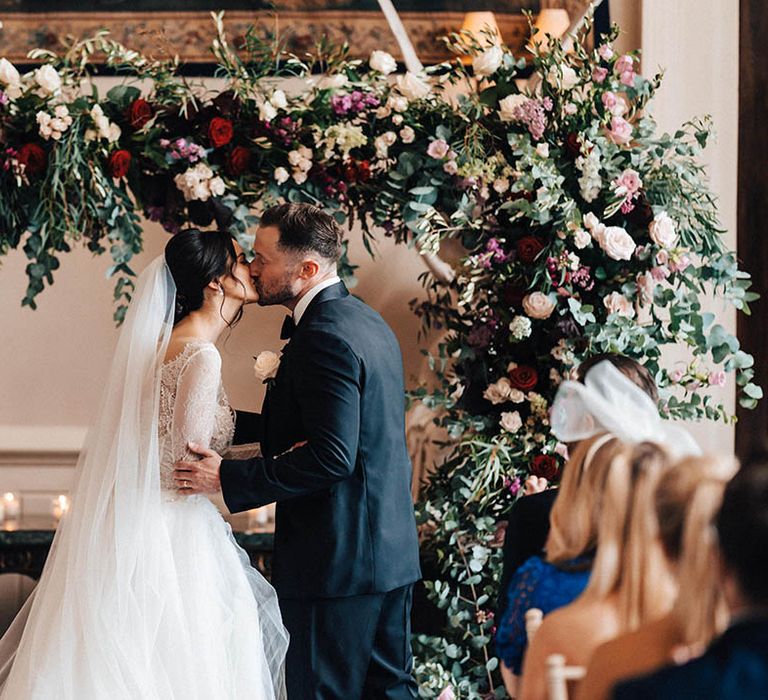 The pink, red, and white wedding flower arch decorates the altar as the bride and groom share their first kiss as a married couple