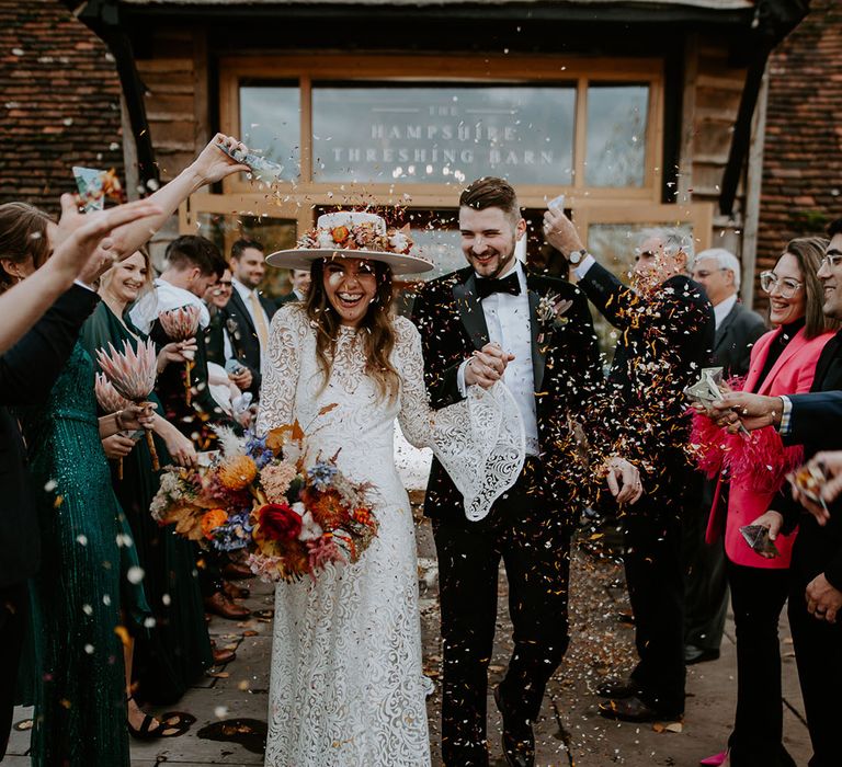 Bride in lace wedding dress with colourful bouquet and white hat with the same colourful flowers has a confetti exit with the groom 