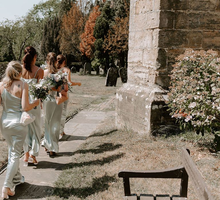 Bridesmaids walk in a row in satin sage green dresses with bouquets to the church for the ceremony 