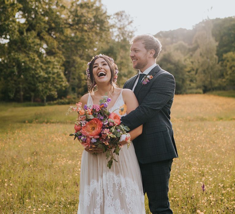 Bride holds colourful floral bouquet filled with peonies as she stands with her groom during golden hour portraits 