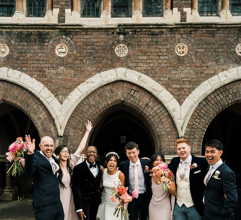 Bride & groom stand beside their wedding party in different styled suits and pastel pink bridesmaid dresses with differing styles whilst holding bright floral bouquets