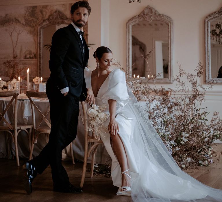 Groom in classic black tux and bowtie and bride in v neck puff sleeve wedding dress with front slit and church length veil sitting in reception room of Eaves Hall wedding venue with chandelier and large dark dried flower arrangements 