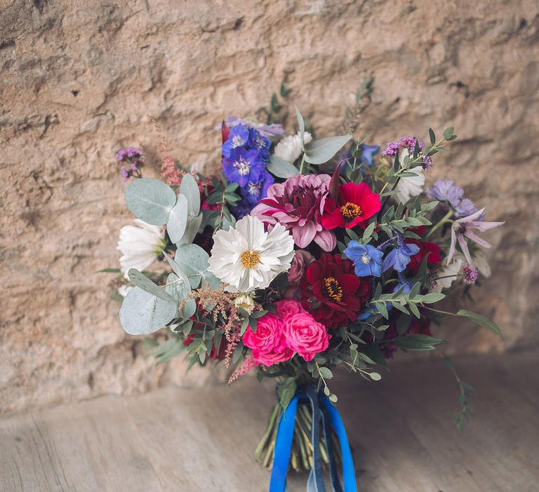 White, pink, red, blue, and purple bridal bouquet tied with blue ribbon and foliage 