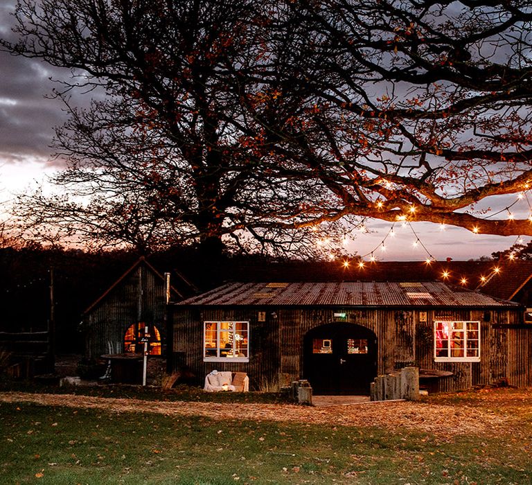 The exterior of Oak Tree Barn wedding venue in Kent at nightfall with fairy lights woven in the trees 