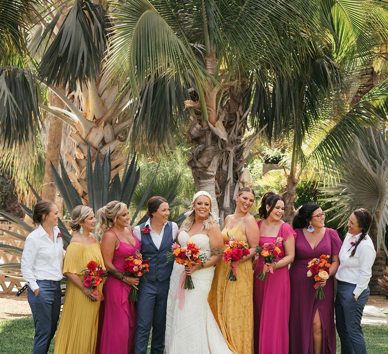 Brides stand with their wedding party in colourful mismatched bridesmaid dresses and white shirts with blue trousers 