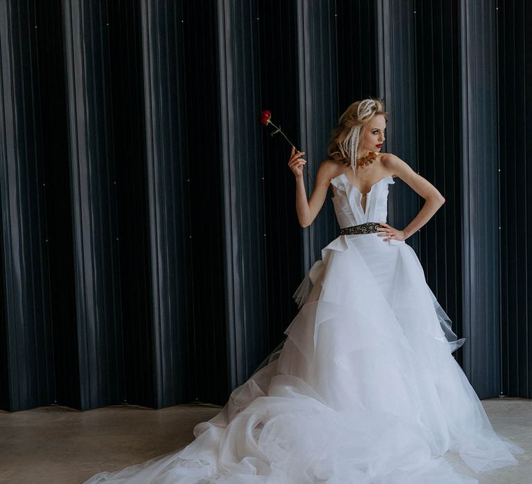 Bride holding a single red rose wearing romantic organza and tulle a-line wedding day bridal ballgown with a flowing skirt, sculpted bandeau top and patterned black belt with dramatic, chunky brass necklace 