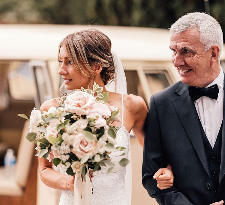 The bride holds a pink rose wedding bouquet walking arm in arm with the father of the bride in a traditional black tuxedo with a white rose buttonhole 