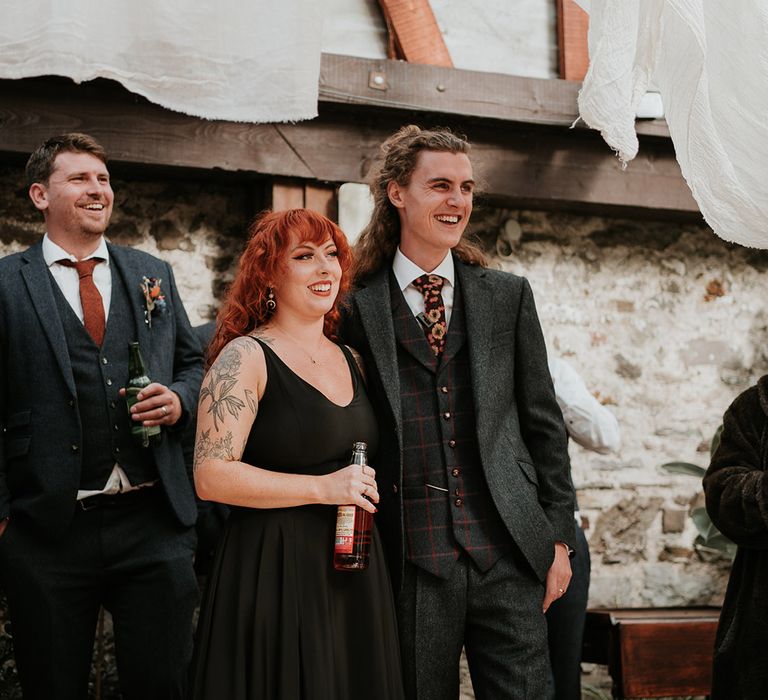 Bride in strappy black dress for wedding in Devon, standing with groom in dark grey three piece suit and burgundy flower patterned tie listening to wedding speeches 