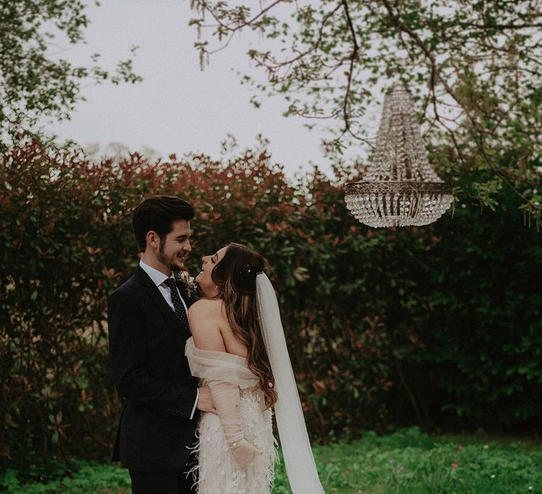 Romantic wedding with the bride in an off the shoulder feather mermaid wedding dress laughing with the groom next to chandelier 