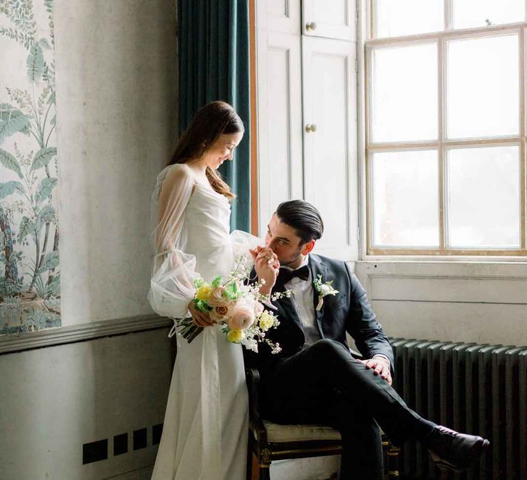 Couple's wedding portraits - groom wearing classic black tuxedo, black bowtie and boutonniere standing with bride in cowl neck satin wedding dress with sheer puff sleeves holding classic spring floral bouquet 