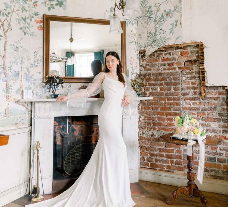 Bride standing by the open fireplace at St Giles House with exposed brickwork and classic wallpaper, wearing cowl neck satin wedding dress with sheer puff sleeves 