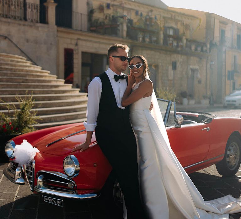 Black bride and groom in sunglasses standing next to their red wedding car 