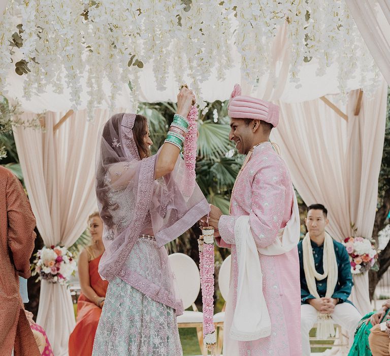 Bride in mint green and dusky pink ombre embellished lehenga and baby pink flower garland performing Indian wedding ritual with groom in light pink sherwani, light pink turban and cream trousers