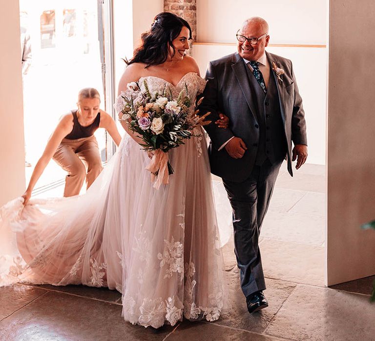 Bride in delicate off the shoulder sweetheart lace wedding dress walking in with the father of the bride in a grey wedding suit 