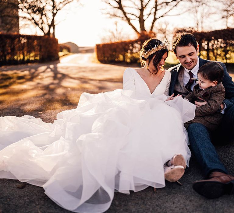 Bride in ruffle gown wearing bride crown sitting with groom in navy suit and their little boy in brown suit 