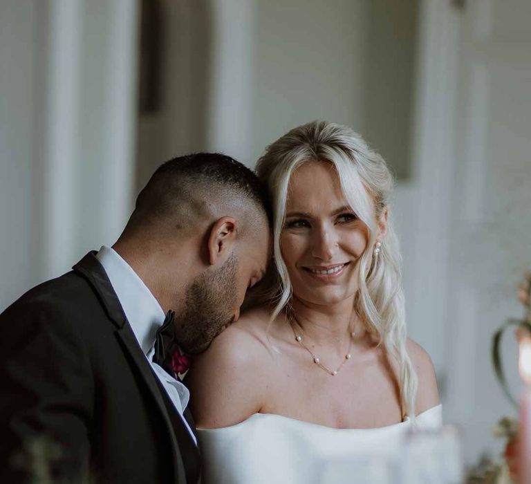 Bride in off the shoulder lace wedding dress and groom in classic three piece grooms tuxedo with colourful spring boutonniere sat at spring wedding tablescape at Frensham Hall wedding venue 