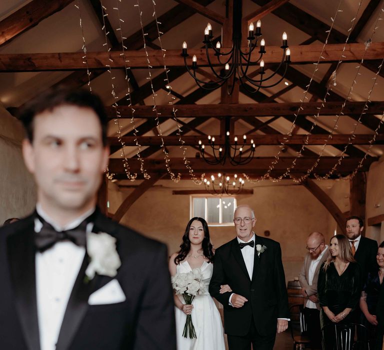 Groom stands at the front of the aisle wearing a black tuxedo whilst the father of the bride walks her down the aisle at Upton barn wedding venue