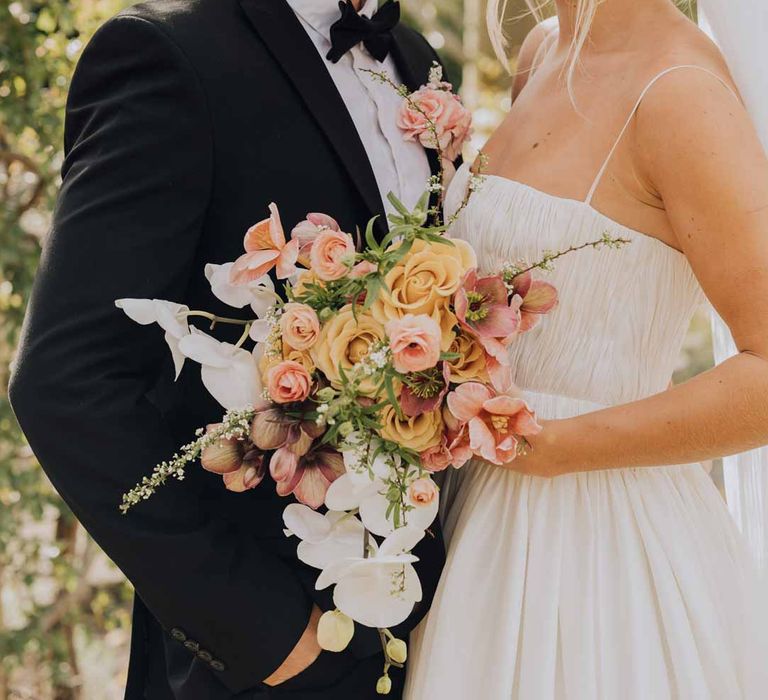 Bride in Watters strappy sleeveless princess wedding dress and chapel length wedding veil holding blush toned bridal bouquet with poppies, ranunculus, roses, tulips and foliage standing with groom in classic grooms tuxedo and bowtie 