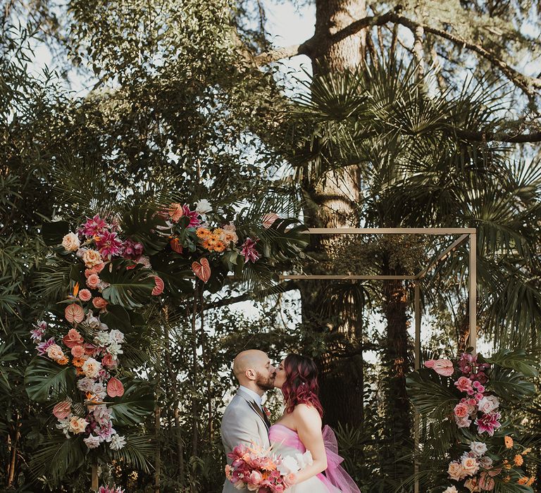 bride in a layered tulle princess wedding dress with pink bow kissing her husband in a light grey wedding suit but their pink tropical flower arch at outdoor Italian villa wedding venue