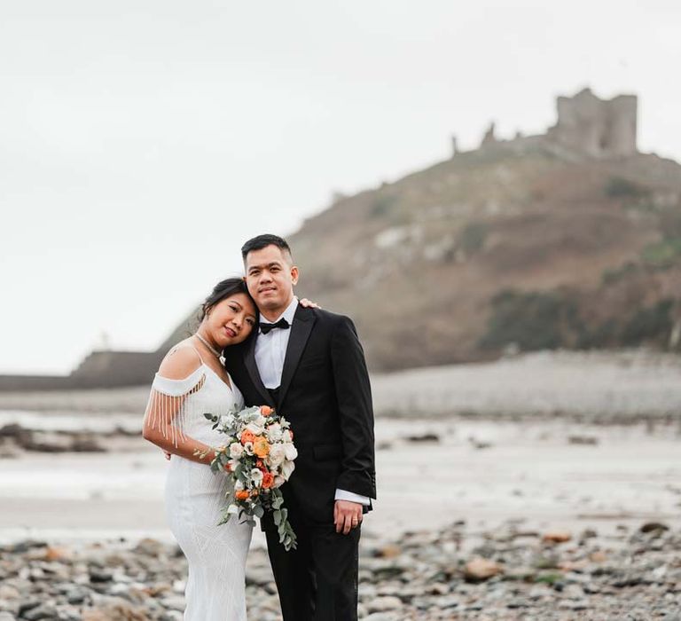 Groom in classic black tux standing with bride in white beaded and sequined slip wedding dress on Welsh beach for Burmese wedding