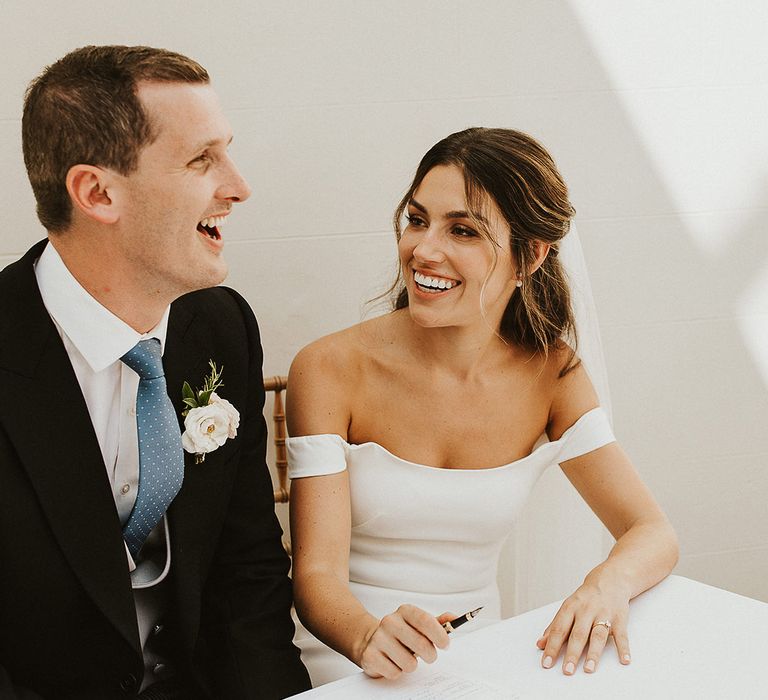 The bride and groom smile brightly at each other as they sign the wedding register