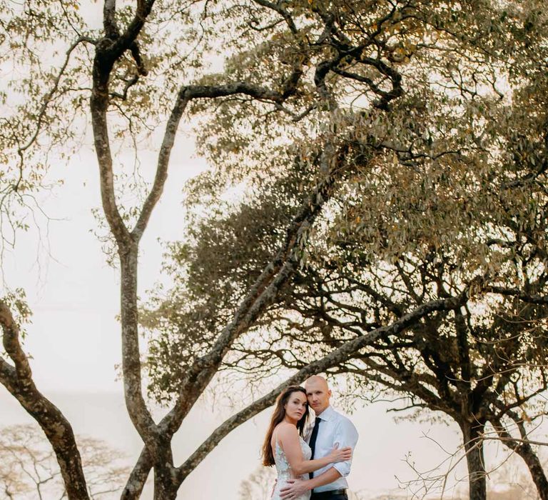 Groom in white shirt, black tie and black suit trousers embracing bride in lace sleeveless wedding dress with puddle train standing by the pool at Giraffe Manor wedding 