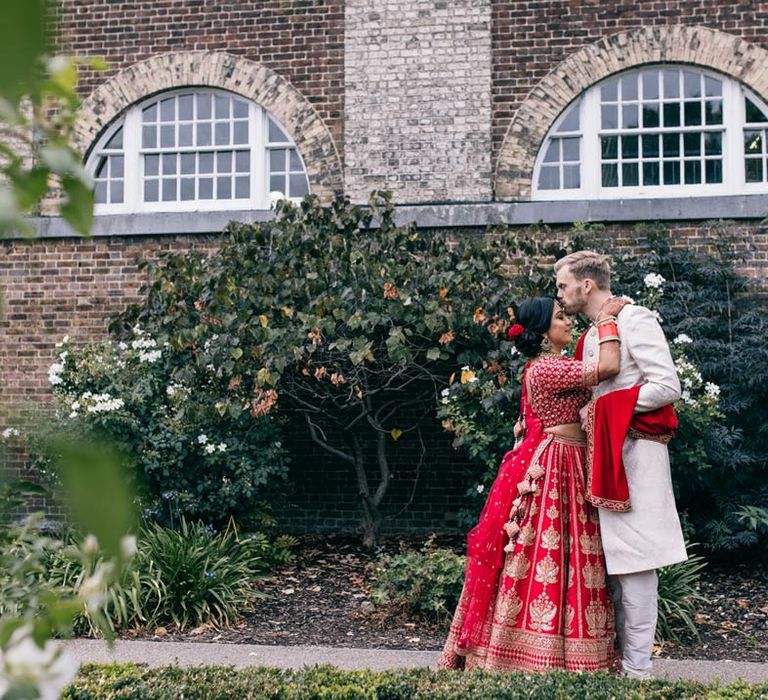 Groom in white and gold sherwani giving bride a kiss on the forehead wearing red and gold lehenga 