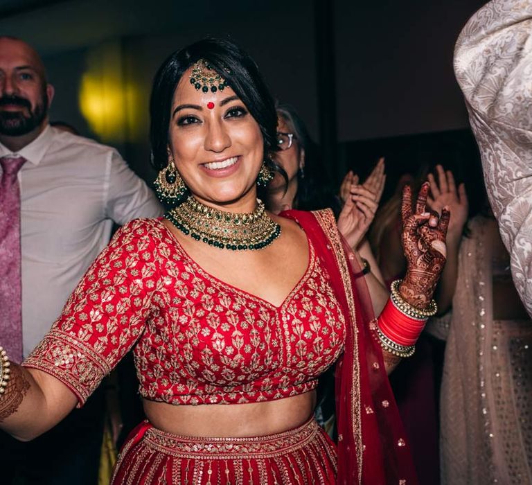 Bride in red and gold lehenga and gold and forest green bridal jewellery dancing at Indian-English fusion wedding 