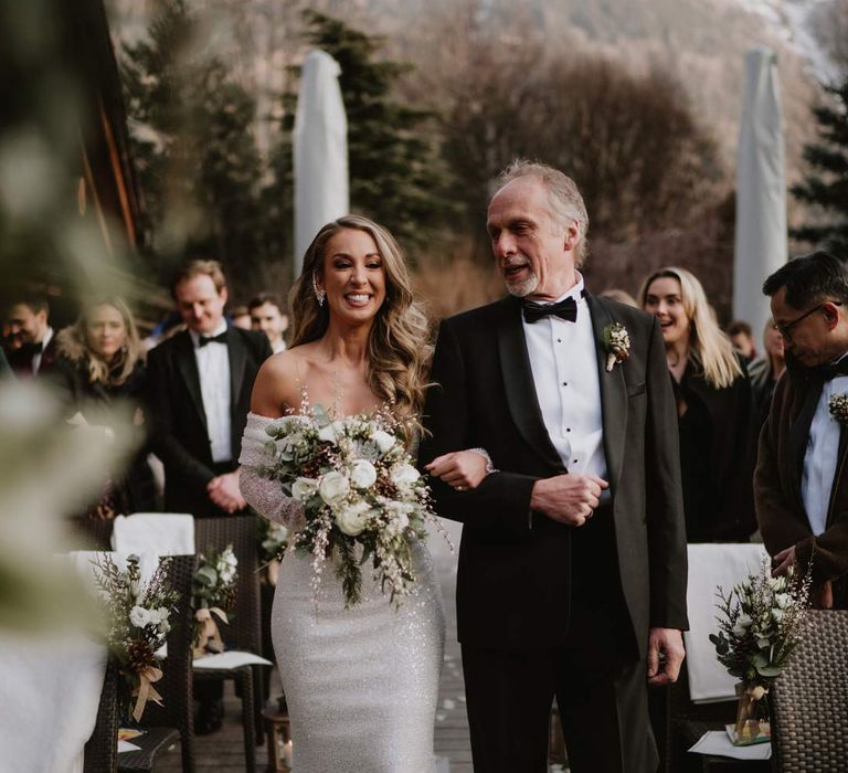 Bride in off shoulder sparkly mermaid wedding dress holding white garden rose, eucalyptus, baby's-breath and foliage bridal bouquet walking down the aisle with father of the bride in classic black tuxedo 