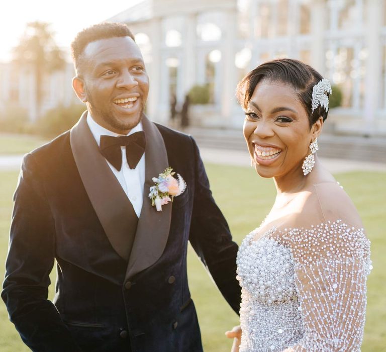 Groom in velvet midnight blue double breasted groom’s blazer, velvet bowtie and peach garden rose boutonniere with bride in illusion v-neck sheer off shoulder long sleeve pearl embellished wedding gown at Syon Park vow renewal