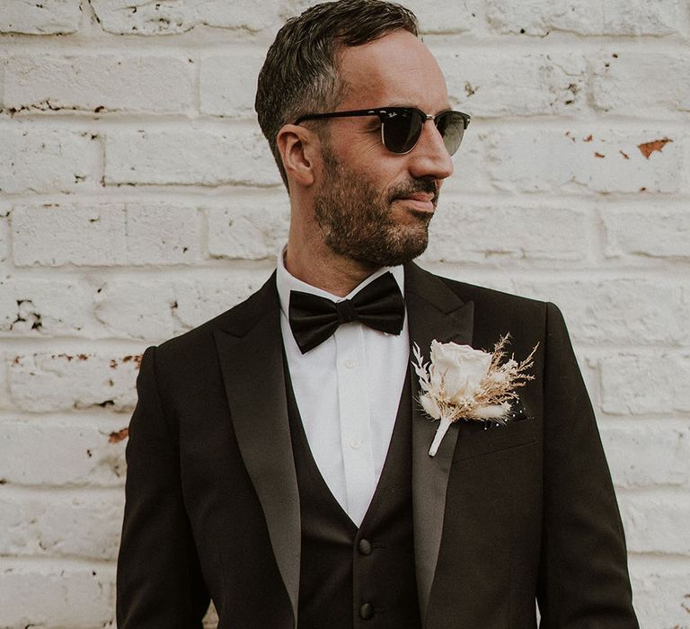 Groom in black tuxedo with black bow tie wearing dried wedding flower buttonhole and black sunglasses 