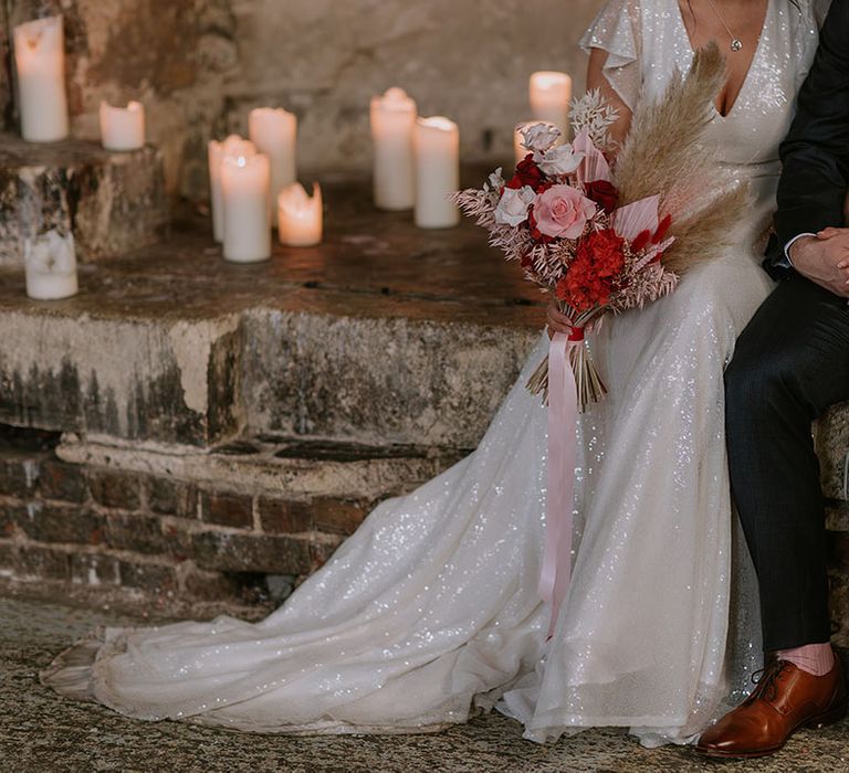 Groom in navy wedding suit with the bride in a sparkly wedding dress carrying a pink and red preserved flower bouquet 