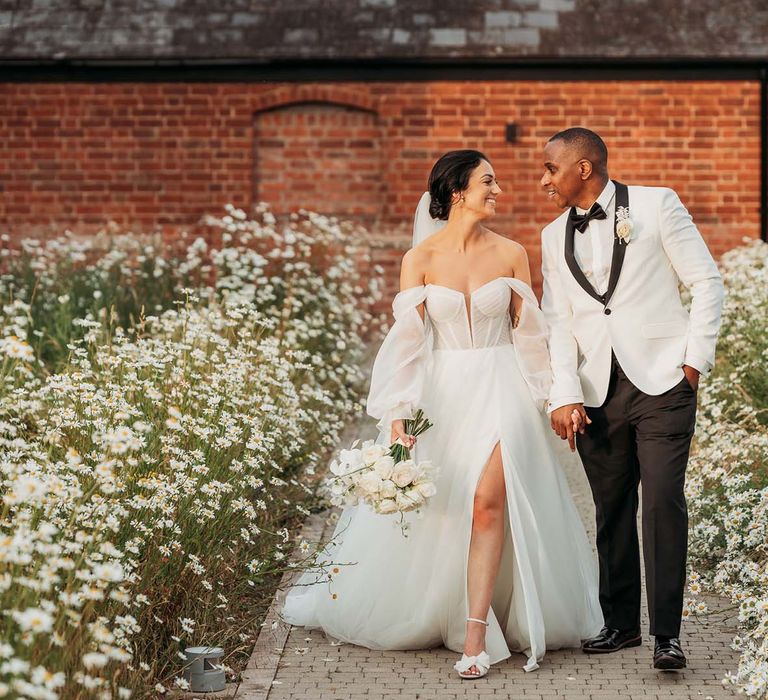 Bride in an off-the-shoulder wedding dress with front slit walks through daisy garden with groom in black-tie tuxedo