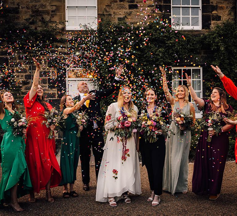 Bridesmaids wearing mismatched bright and colourful dresses throw confetti over the brides 