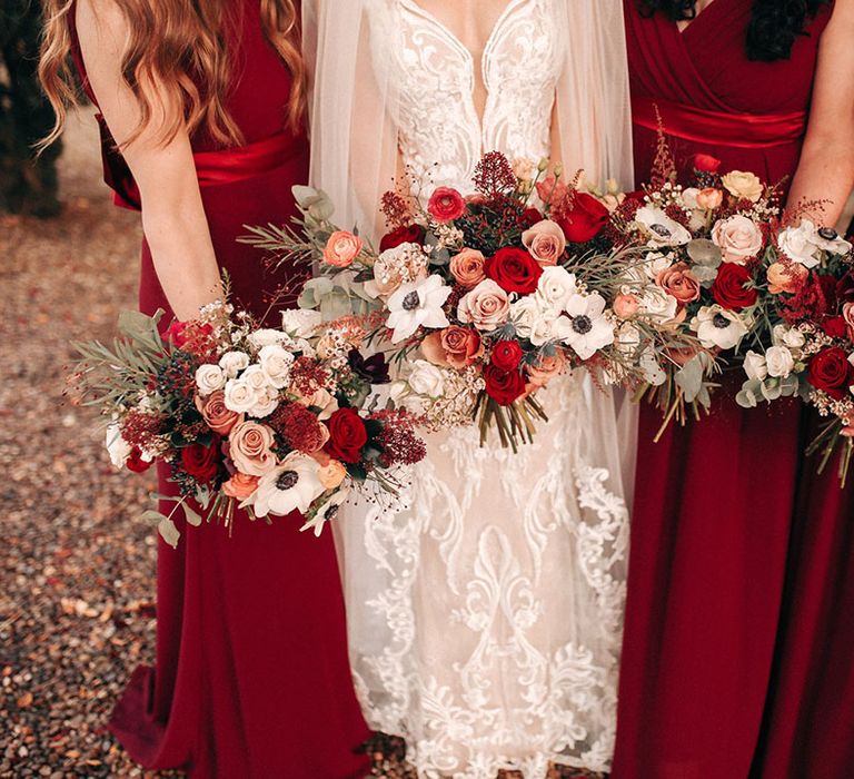 Bridal party wearing red bridesmaid dresses with the bride holding red rose and white anemone bouquet 