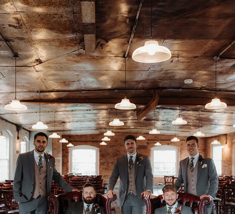 The groom and groomsmen pose together for a group photo with the groomsmen posing on red leather chairs 