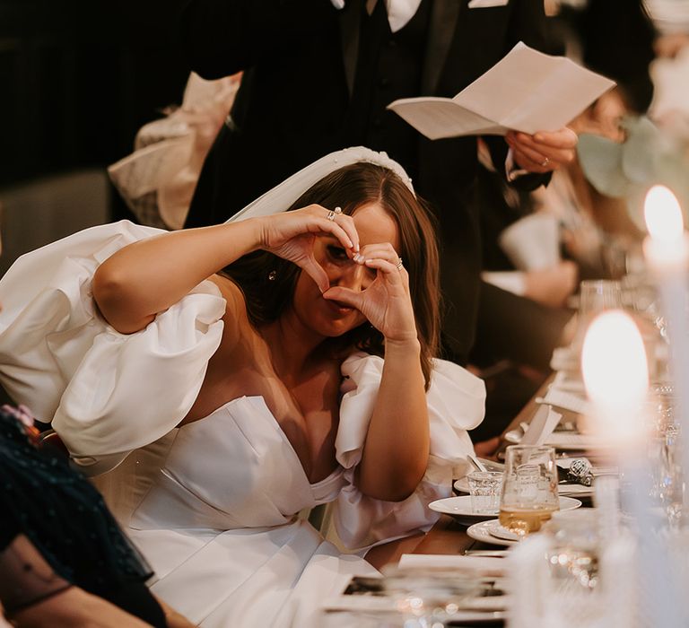 Bride makes heart shape with her hands at the camera while groom reads his wedding speech 