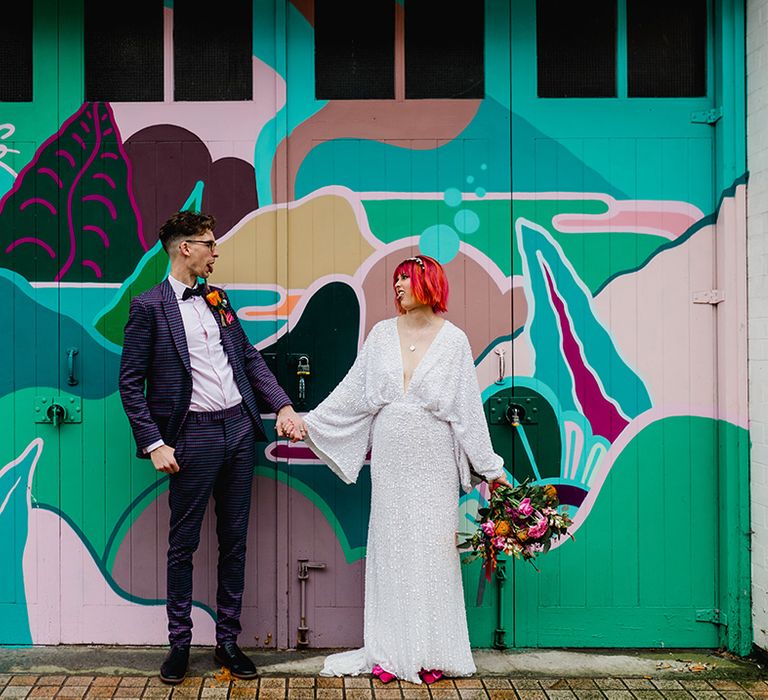 Bride in bat wing sequin wedding dress with groom in purple checkered suit posing in front of colourful painted room 