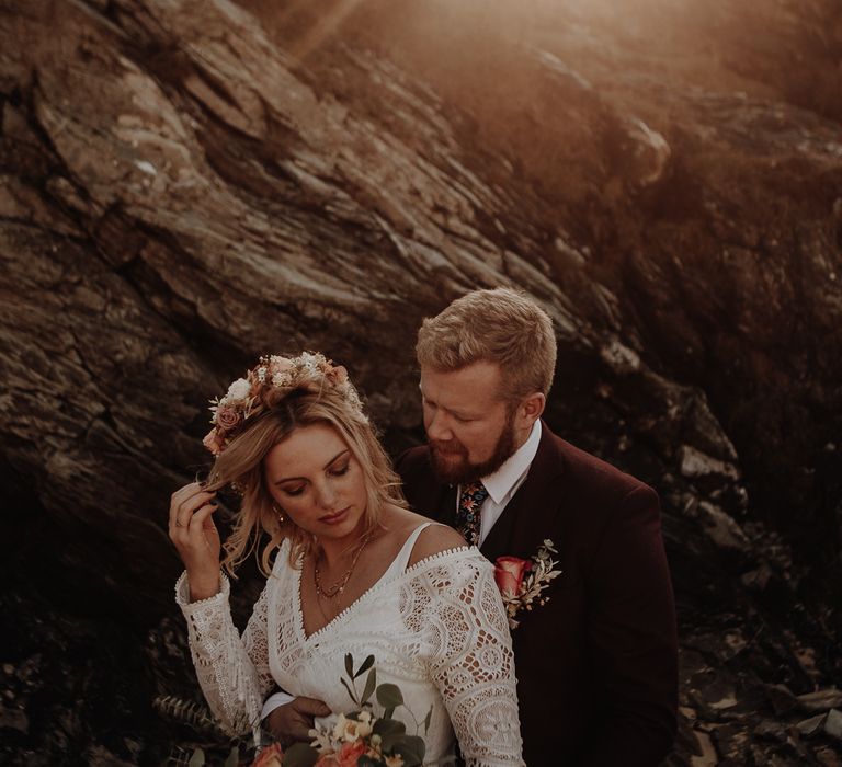 Groom with rose boutonnière and burgundy suit embraces with bride in boho style wedding dress