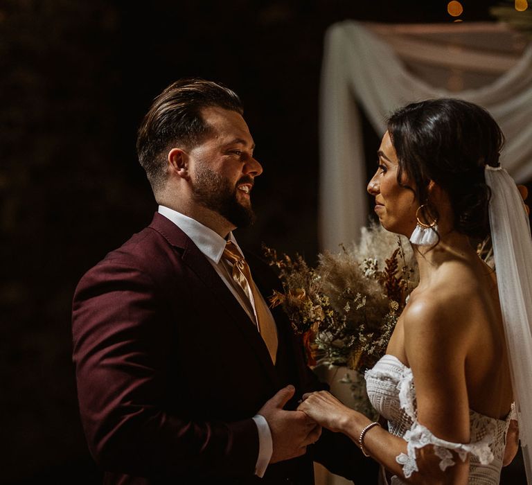 Groom and emotional bride hold hands by wedding arch