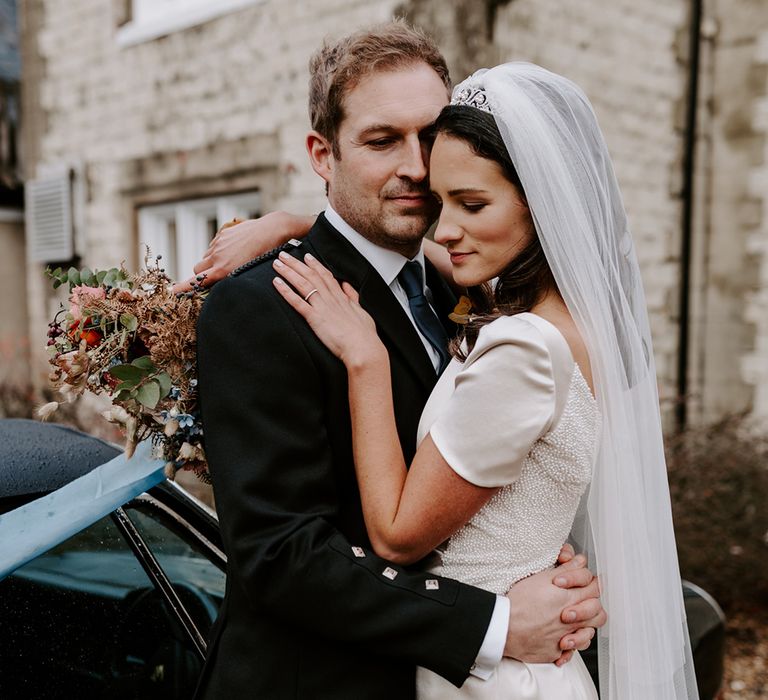 Bride and groom embrace next to their wedding car. The bride wears an embellished white gown and the groom wears a kilt.