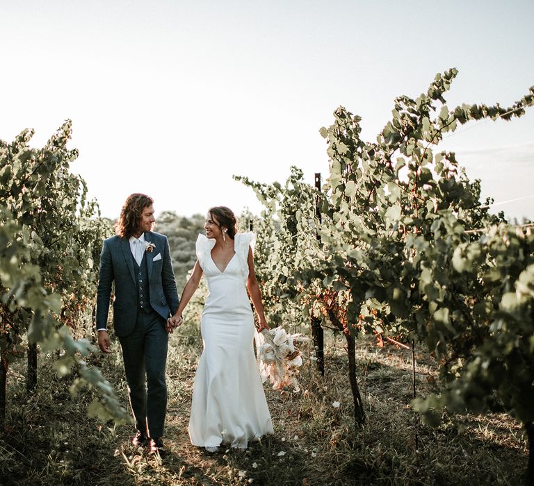 Groom in three-piece navy suit holding his brides hand in a plunging neckline Rime Arodaky wedding dress with ruffle sleeves 