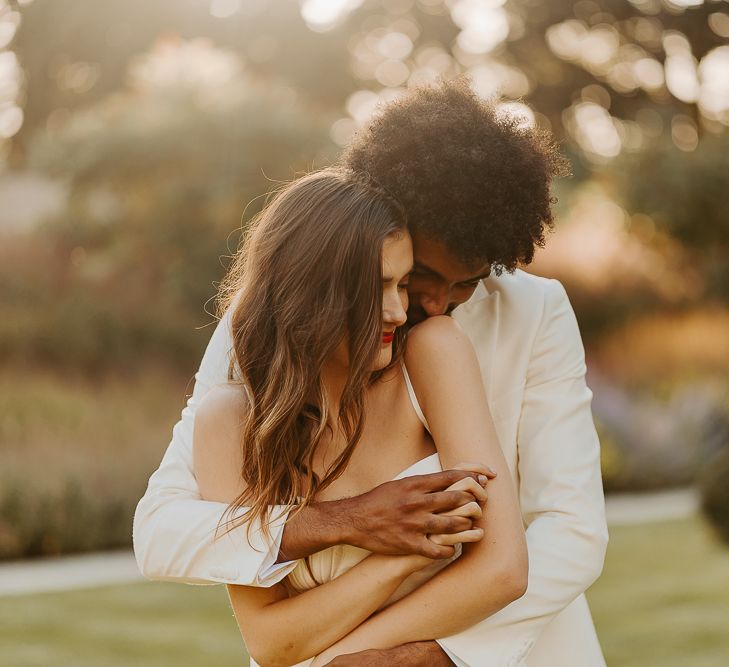 Golden hour portrait with Black groom with afro hair and white tuxedo jacket embracing his bride with long brown hair and red lipstick