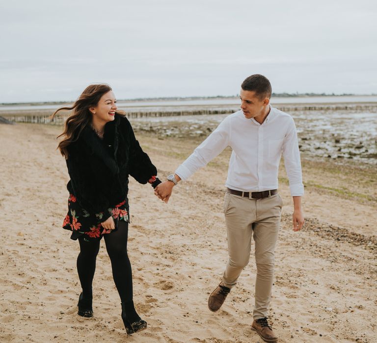 Bride-to-be in a black faux fur coat on the beach holding hands with her husband-to-be during their engagement photo shoot with Grace Elizabeth 