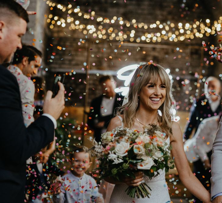 White bride with fringe holding a bouquet with groom in grey jacket under a shower of confetti